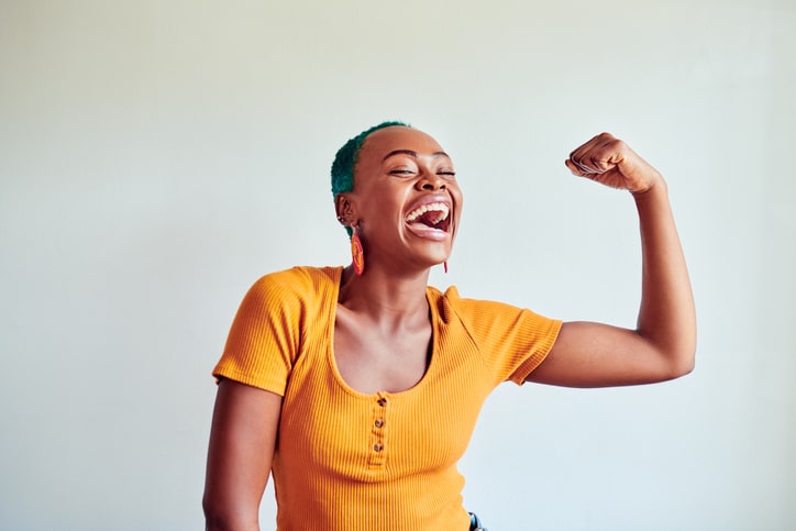 A young woman in a yellow top and with a shaved head flexes her forearm and smiles widely